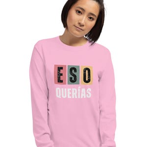 May include: A light pink long-sleeve shirt with the text "ESO QUERÍAS" in a colorful block design. The word "ESO" is in a pink block, "QUERÍAS" is in a white font.