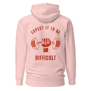 Puede incluir: Sudadera con capucha rosa claro con un gráfico rojo de una mano sosteniendo una barra con pesas, con las palabras "EXPECT IT TO BE DIFFICULT" impresas arriba y abajo. La sudadera tiene capucha y mangas largas.