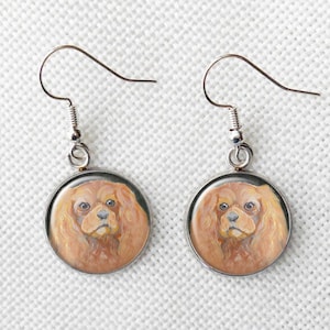 May include: A pair of silver dangle earrings with round, silver-rimmed, glass cabochons featuring a painted image of a Cavalier King Charles Spaniel dog with a light brown and white coat.