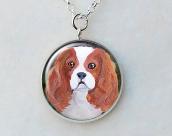May include: A silver-toned pendant necklace featuring a round charm with a painted portrait of a Cavalier King Charles Spaniel dog. The dog has brown and white fur and a sweet expression.