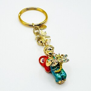 May include: A gold-toned metal keychain featuring a small figure with red wings and teal clothing, holding a dragonfly charm adorned with multicolored crystals. The keychain includes a circular ring and a small dangling dragonfly accent piece.