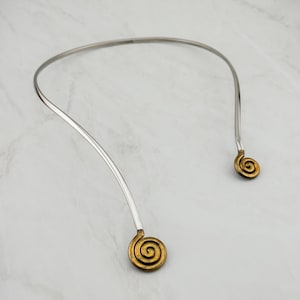 Könnte beinhalten: Ein offenes Halsband in Silberoptik mit einem glatten, geschwungenen Band, das an beiden Enden in einem kreisförmigen, goldfarbenen Spiraldesign endet. Das Metallstück ist starr und behält eine breite, abgerundete Form bei, wobei es einen Kontrast zwischen der polierten silbernen Oberfläche des Hauptbandes und der strukturierten, antik wirkenden goldenen Oberfläche der Spiralenden aufweist.