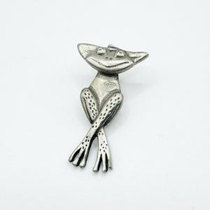 May include: A silver-toned metal brooch shaped like a frog with a wide head, pointed ears, and a smiling expression. The frog has long, slender legs crossed over each other, with visible textured spots on the lower limbs and a smooth torso.