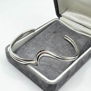 May include: A silver-toned cuff bracelet featuring a double-band design that curves into a central wave-like peak. The metal has a polished, reflective finish, and the open-ended structure allows for adjustment around the wrist.