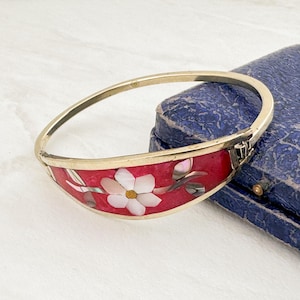 May include: A hinged bangle bracelet featuring a curved metal band with a vibrant red inlay. The inlay is decorated with a white floral motif made of iridescent shell pieces, including a central five-petaled flower and green leaf accents.
