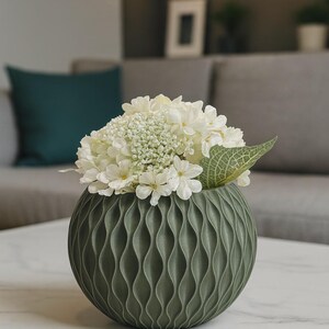 Può includere: Un vaso decorativo verde oliva con un motivo a onde, pieno di fiori bianchi e verde. Il vaso rotondo è posto su un tavolo di marmo bianco, creando un'estetica moderna e minimalista. Il vaso ha un diametro di circa 20 cm.