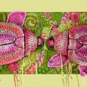 Könnte beinhalten: Ein lebendiges, handgefertigtes Häkelkunstwerk mit zwei großen, stilisierten insektenartigen Formen in Rosa-, Orange- und Grüntönen. Das Kunstwerk enthält florale Elemente und einen grünen Hintergrund, wodurch ein skurriles, von der Natur inspiriertes Design entsteht.