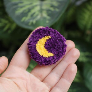 May include: A handmade, round, purple crochet patch featuring a yellow crescent moon design. The patch is held in a hand, with a blurred green background of foliage. The patch is a decorative item.