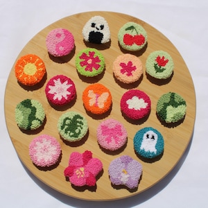 Pode incluir: Uma coleção de patches bordados redondos e coloridos com vários desenhos, incluindo flores, frutas e um fantasma, dispostos numa bandeja de madeira. Os patches apresentam cores vibrantes e costuras detalhadas, criando um sortido visualmente apelativo.