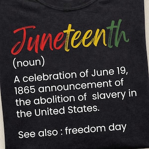May include: Black t-shirt with the word "Juneteenth" in red, yellow, and green script. Below, the shirt describes Juneteenth as a celebration of the 1865 announcement of the abolition of slavery in the United States. The text also includes "See also: freedom day."