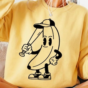 Könnte beinhalten: Gelbes Sweatshirt mit einer Cartoon-Bananenfigur, die eine Baseballkappe trägt und einen Schläger hält. Die Banane hat ein lächelndes Gesicht und ist mit schwarz-weißen Handschuhen und Schuhen bekleidet. Das Design ist in schwarzer Tinte.