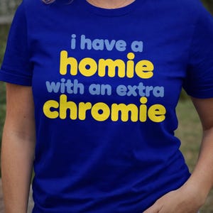 Può includere: T-shirt blu reale con la scritta "i have a homie with an extra chromie" in giallo e azzurro. La maglietta è girocollo e a maniche corte.
