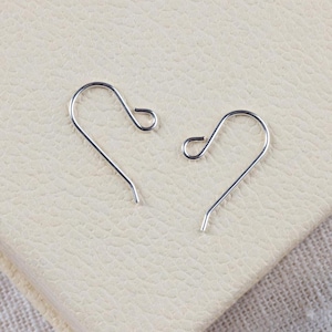 May include: Two silver earring hooks, also known as French hooks, are shown on a light beige surface. The hooks are made of metal and have a simple, elegant design.