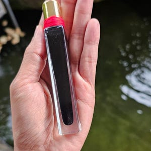 May include: A rectangular perfume bottle with a gold cap and a red band. The bottle is clear with a dark, opaque liquid inside. The bottle is held in a hand against a blurred background of greenery and water.