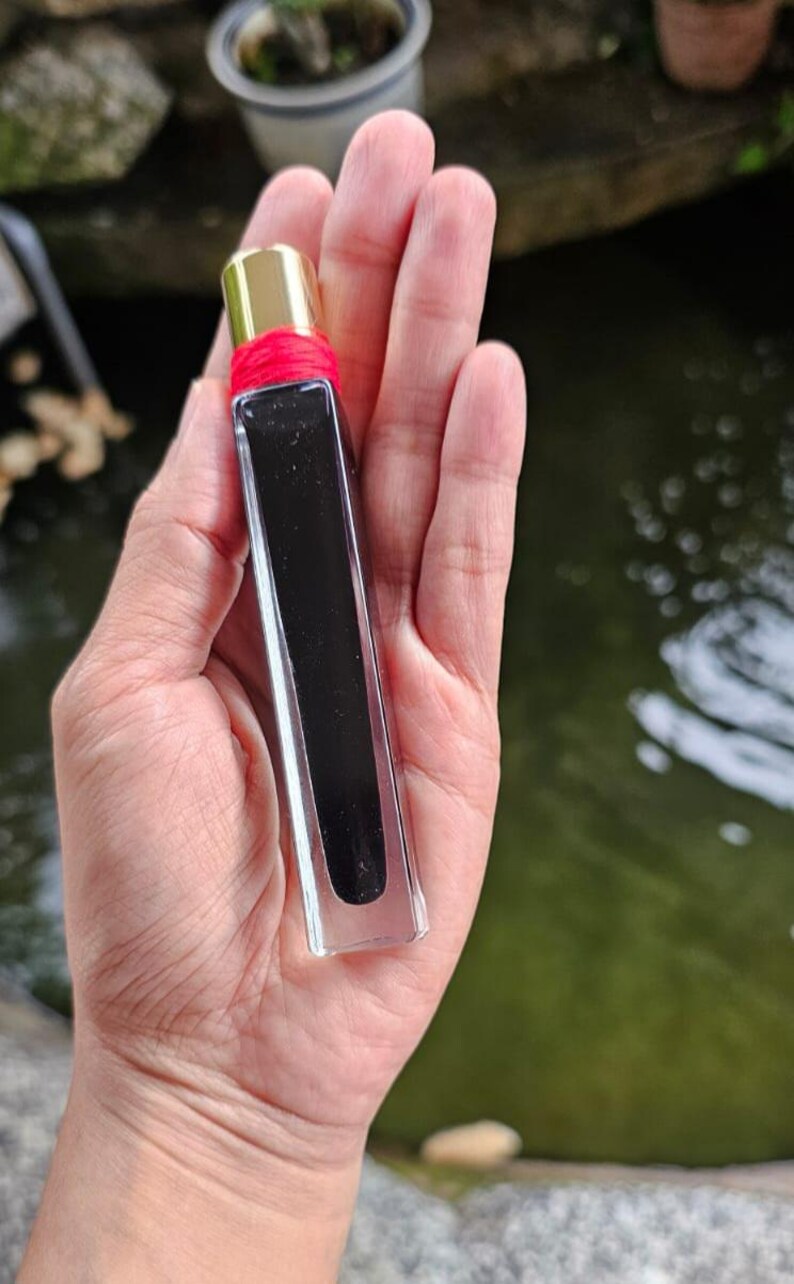 May include: A rectangular perfume bottle with a gold cap and a red band. The bottle is clear with a dark, opaque liquid inside. The bottle is held in a hand against a blurred background of greenery and water.