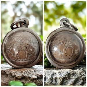 May include: Two antique, round, silver-toned pendants with intricate engraved designs. Each pendant features a central motif of what appears to be a fruit or vessel, surrounded by detailed patterns. The pendants are displayed on a natural stone surface.