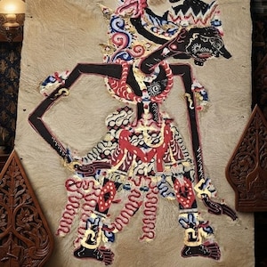 Pode incluir: Um fantoche de sombra Wayang Kulit com uma figura preta, vermelha e dourada com padrões intrincados sobre um fundo de pele castanho-claro texturizado. O texto ZEMystical Shop está impresso a branco na parte inferior do expositor.
