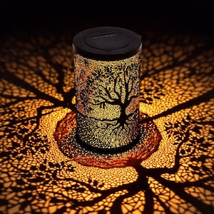 Puede incluir: Una linterna cilíndrica negra con un diseño de árbol de la vida. La linterna proyecta una sombra cálida y detallada del árbol sobre la superficie circundante. La iluminación interior crea un ambiente acogedor y acogedor.