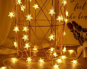 May include: A decorative copper-colored metal basket with a geometric design, adorned with a string of warm white star-shaped fairy lights. The lights are draped around the basket and scattered on the surface, creating a cozy ambiance.