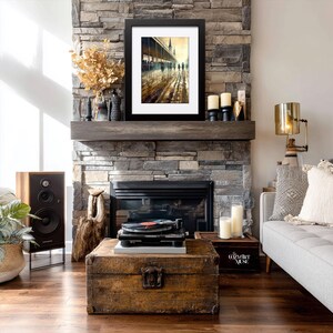May include: A framed art print depicting a rainy street scene with figures walking under a covered walkway. The artwork is displayed above a fireplace with a stone facade. A vintage record player sits on a wooden trunk, with a speaker and lamp nearby.