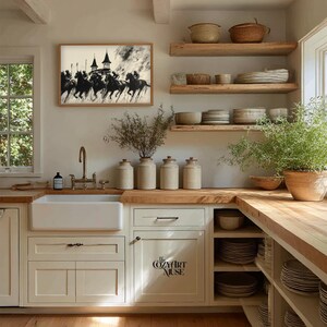 May include: A kitchen with a farmhouse sink, wooden countertops, and open shelving. A framed black and white artwork of riders on horseback hangs above the sink. The cabinet door has the text "The Cozy Art Muse".