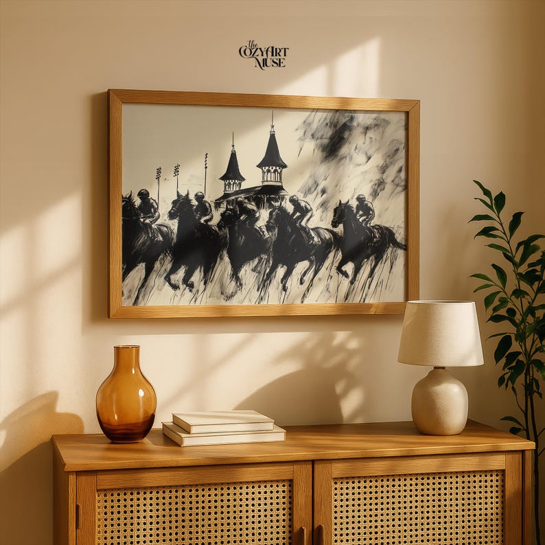 May include: A framed black and white print depicting a horse race with jockeys. The artwork is in a light wood frame and hangs above a wooden cabinet. A decorative amber glass vase and a small lamp are on the cabinet.