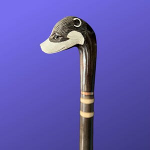 Puede incluir: Bastón hecho a mano con un mango de cabeza de hurón tallada. La cabeza del hurón está detallada con tonos negros, blancos y grises. El eje marrón oscuro tiene bandas decorativas de color marfil. El bastón está sobre un fondo azul.