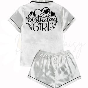 May include: White satin pyjama set with short sleeves and shorts. The top has a black trim and the text "birthday girl" with hearts printed on the back.