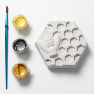 May include: A white hexagonal plaster plaque featuring a raised bee and honeycomb pattern, accompanied by a blue paintbrush and three small circular containers filled with metallic gold and silver paint for decorative crafting projects.