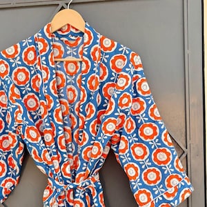 May include: A blue floral patterned robe with orange and white flower designs. The robe is hanging on a wooden hanger against a gray door. The robe has a matching belt.