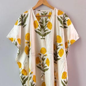 Könnte beinhalten: Weißes Kaftankleid mit V-Ausschnitt und kurzen Ärmeln. Das Kleid hat ein sich wiederholendes Muster aus gelben Blumen mit grünen Stielen und Blättern. Ein Kordelzug schnürt die Taille zusammen. Das Kleidungsstück hängt an einem Holzbügel.