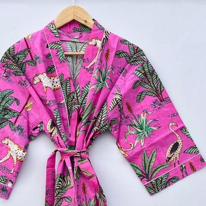 May include: A pink cotton robe with a white trim. The robe features a jungle print with leopards, zebras, palm trees, and flamingos.