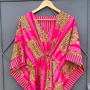 May include: A vibrant pink kaftan with a V-neck and a drawstring waist. The fabric features a bold tiger stripe pattern in shades of yellow and black, set against vertical pink stripes. The sleeves and neckline are trimmed with white stripes.