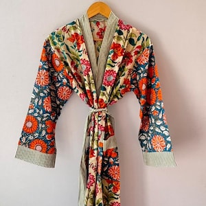 Puede incluir: Bata con estampado floral con fondo crema, con flores naranjas, rojas y turquesas. Las mangas y el ribete tienen rayas verdes y blancas. La bata está colgada de una percha de madera.