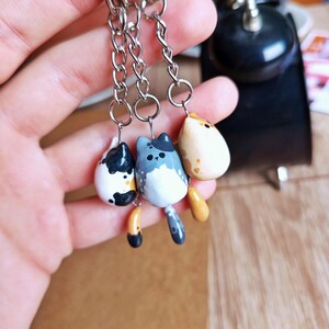 May include: Three cat-shaped keychains with silver chain attachments. The cats are painted in black and white, gray and blue, and beige with orange accents. Each keychain has a small tail. The keychains are held in a hand.