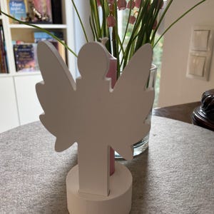 Może przedstawiać: Biała drewniana figurka anioła z rozłożonymi skrzydłami i okrągłą podstawą. Anioł to prosty element dekoracyjny, idealny do dekoracji wnętrz. Figurka stoi na szarej powierzchni.