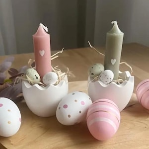Può includere: Due portacandele in ceramica bianca a forma di uovo con candele rosa e verdi. Ogni portacandele è decorato con un piccolo cuore bianco e circondato da piccole uova macchiate. I portacandele sono su una superficie di legno con quattro uova decorative.
