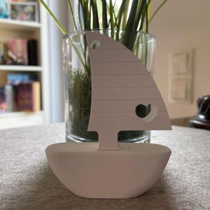 Może przedstawiać: Biały drewniany żaglówka z wycięciem w kształcie serca w żaglu. Łódź stoi na szarej powierzchni, za nią szklany wazon z zielenią. Żaglówka to przedmiot dekoracyjny.