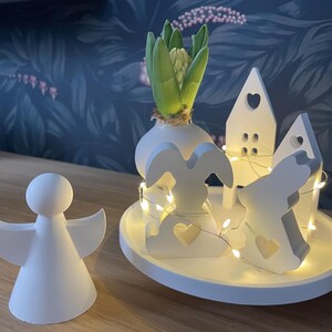 Könnte beinhalten: Eine weiße Keramik-Engelsfigur steht neben einem dekorativen Arrangement. Das Arrangement umfasst eine weiße Vase mit einer Hyazinthe, einen Hasen und zwei hausförmige Objekte, alles auf einem weißen Tablett, beleuchtet von Lichterketten.