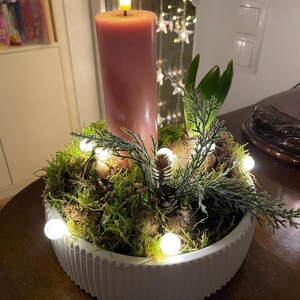 Op de afbeelding: Een brandende, hoge, stoffig roze kaars staat in een witte, geribbelde, ondiepe kom gevuld met groen mos, kleine dennenappels en takjes groen. Kleine, ronde, witte lampjes zijn door de opstelling verspreid en creëren een warme, feestelijke sfeer.