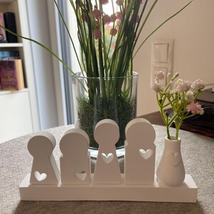Peut inclure: Un ensemble de figurines familiales en bois blanc avec des découpes en forme de cœur. L'ensemble comprend quatre figurines et un petit vase avec un motif de cœur, contenant des fleurs roses. Un vase en verre avec des tiges vertes et des fleurs roses est en arrière-plan.