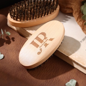 Puede incluir: Dos cepillos de barba de madera con cerdas oscuras. Uno de los cepillos es de forma ovalada con las iniciales "JR" y el nombre "Jeremiah" grabados en el mango de madera. El otro cepillo, en el fondo, también es de madera, con cerdas oscuras.