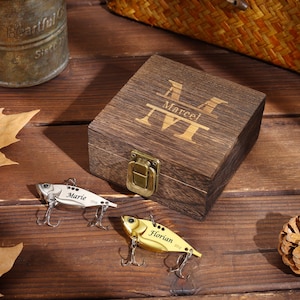 May include: A wooden box with the name "Marcel" engraved on the lid, next to two fishing lures. One lure is silver and says "Marie", the other is gold and says "Florian". The box has a metal clasp.