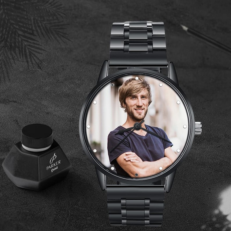 Op de afbeelding: Zwarte polshorloge met een foto op de wijzerplaat. Het horloge heeft een zwarte metalen band en een zwarte kast. Een zwarte inktfles met "Parker Quink" staat naast het horloge.