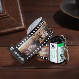 May include: A novelty keychain featuring a miniature film canister with a green and black label that reads "X-TRA 400" and "FUJIFILM". A strip of film with printed images is attached. The film strip has a musical note and sound wave graphic.