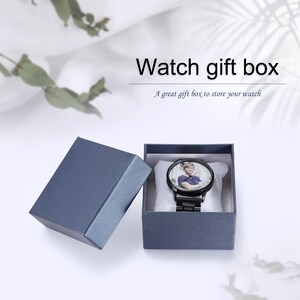 Op de afbeelding: Een blauwe horlogegeschenkdoos met een zwart horloge erin. Het horloge heeft een foto op de wijzerplaat. De doos is open en onthult het horloge op een wit kussen. Tekst op de afbeelding luidt "Watch gift box".