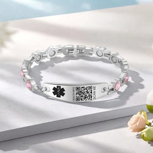 May include: A silver medical ID bracelet with pink heart-shaped accents. The bracelet features a central plate with a medical symbol, the words "HEALTH INFO" and a QR code. The bracelet is designed to be worn on the wrist.