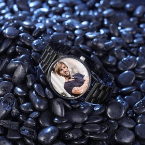 Op de afbeelding: Een zwart polshorloge met een roestvrijstalen band en een ronde wijzerplaat. De wijzerplaat toont een kleurenfoto van een persoon. Het horloge ligt op een bed van donkerblauwe stenen.