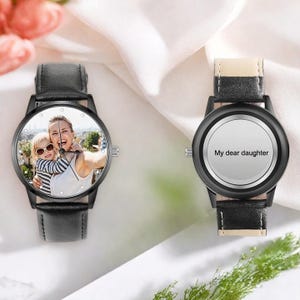 Puede incluir: Dos relojes de pulsera negros. Una esfera muestra una foto de una mujer y un ni&ntilde;o. La otra esfera es blanca con el texto "My dear daughter". Ambos relojes tienen correas de cuero negro y cajas negras.