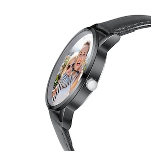 Puede incluir: Un reloj de pulsera negro con una correa de cuero gris. La esfera del reloj muestra una fotograf&iacute;a en color de dos personas. El reloj tiene un bisel negro y una corona plateada. La correa mide aproximadamente 2 cm de ancho.